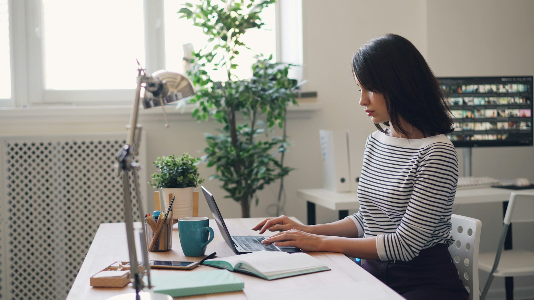 A woman working at a desk with a laptop, beside a window and a plant